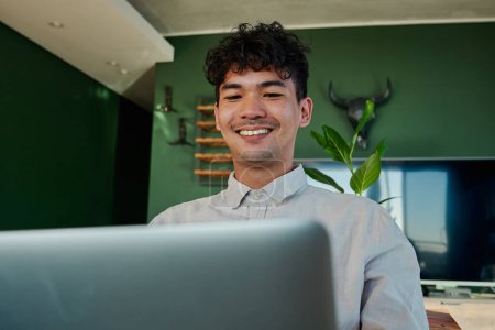 Téléchargez les photos : Foyer sélectif de jeune homme multiracial portant chemise souriant tout en utilisant un ordinateur portable dans le salon à la maison - en image libre de droit