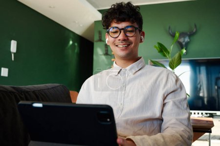 Junger multiethnischer Mann im Hemd lächelt, während er zu Hause auf dem Sofa ein digitales Tablet mit drahtlosen Kopfhörern benutzt
