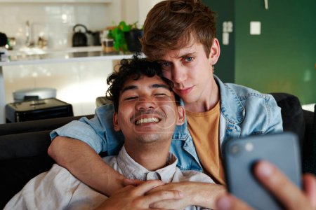 Foto de Joven pareja gay tomando selfie con teléfono móvil mientras se abraza en el sofá en la sala de estar en casa - Imagen libre de derechos