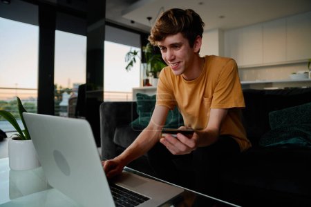 Foto de Feliz joven caucásico hombre con camiseta que sostiene la tarjeta de crédito mientras hace compras en línea en el ordenador portátil en casa - Imagen libre de derechos