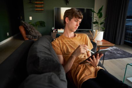 Foto de Joven hombre caucásico usando camiseta escribiendo con bolígrafo digitalizado en tableta digital en la sala de estar en casa - Imagen libre de derechos