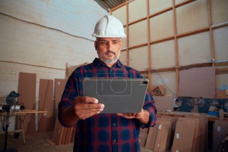 Photo for Focused multiracial mid adult man in safety helmet using digital tablet by woodpile in woodworking factory - Royalty Free Image