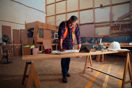 Foto de Multiracial hombre adulto medio que trabaja junto a la tableta digital y herramientas en la mesa en la fábrica de carpintería - Imagen libre de derechos