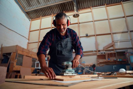 Photo pour Multiracial mi-adulte homme souriant tout en portant des cache-oreilles et en utilisant un outil électrique sur le bois dans l'usine de travail du bois - image libre de droit