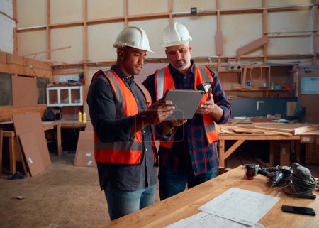 Photo pour Hommes multiraciaux en gilet réfléchissant regardant tablette numérique près des outils électriques et du papier dans l'usine de travail du bois - image libre de droit