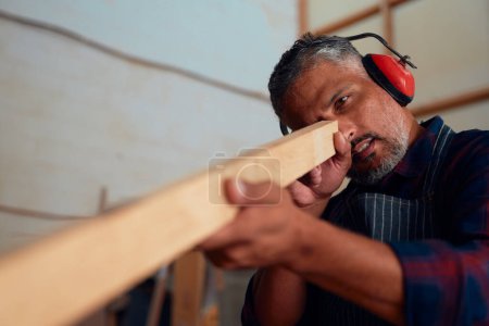 Photo pour Homme adulte moyen multiracial portant des cache-oreilles tenant des planches de bois dans une usine de travail du bois - image libre de droit