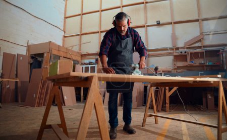 Photo pour Homme adulte moyen multiracial portant un tablier et des cache-oreilles et utilisant un outil électrique sur bois dans une usine de travail du bois - image libre de droit