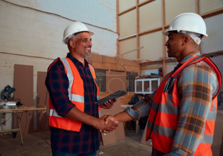 Téléchargez les photos : Hommes multiraciaux portant des vêtements de travail protecteurs souriant tout en serrant la main à l'usine de travail du bois - en image libre de droit