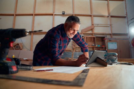 Photo pour Multiracial mi-homme adulte écriture avec stylo numérisé sur tablette numérique à côté des outils électriques dans l'usine de travail du bois - image libre de droit