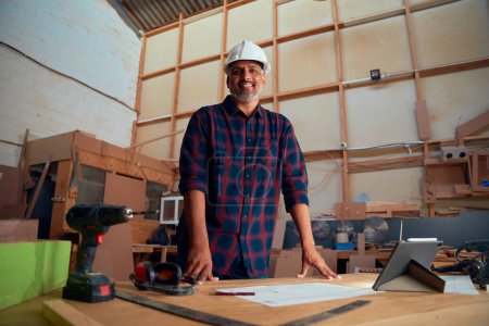 Foto de Multiracial hombre adulto medio en casco de seguridad sonriendo cerca de tableta digital y herramientas eléctricas en la fábrica de carpintería - Imagen libre de derechos