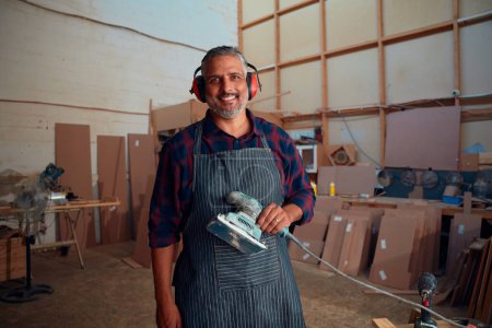 Téléchargez les photos : Multiracial mi-adulte homme souriant tout en portant des cache-oreilles et tenant outil électrique dans l'usine de travail du bois - en image libre de droit
