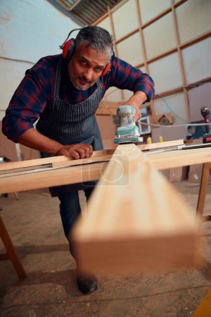 Photo pour Homme adulte moyen multiracial portant des cache-oreilles à l'aide d'un outil électrique sur une planche de bois dans une usine de travail du bois - image libre de droit