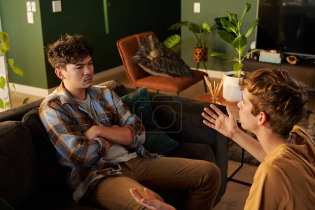 Foto de Frustrada joven pareja gay en ropa casual discutiendo y haciendo gestos en la sala de estar en casa - Imagen libre de derechos