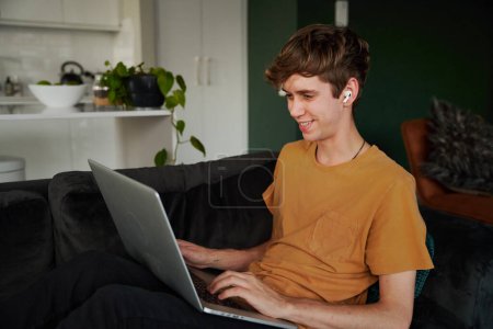 Photo for Happy young caucasian man wearing t-shirt working on laptop with wireless headphones on sofa at home - Royalty Free Image