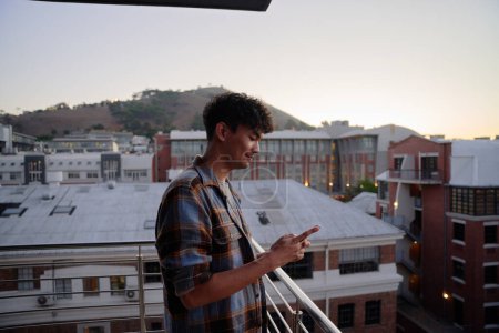 Téléchargez les photos : Vue du profil de jeune homme multiracial en chemise utilisant le téléphone sur le balcon de l'appartement pendant le coucher du soleil - en image libre de droit