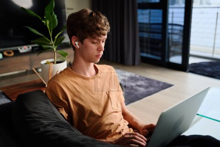 Téléchargez les photos : Focalisé jeune homme caucasien portant t-shirt de travail sur ordinateur portable avec casque sans fil sur canapé à la maison - en image libre de droit