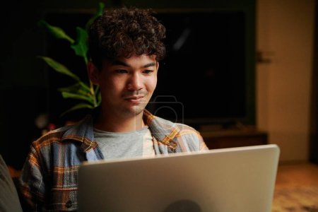 Photo for Cheerful young multiracial man wearing checkered shirt looking away while using laptop on sofa in house - Royalty Free Image