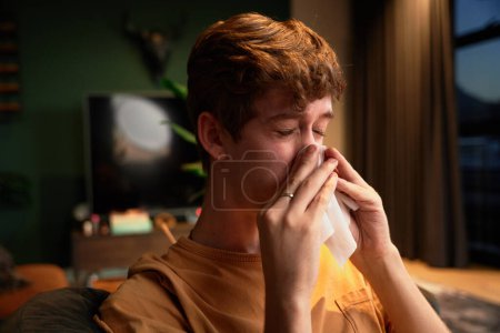 Foto de Enfermo joven caucásico hombre con camiseta que sopla la nariz junto a la computadora portátil en el sofá en la sala de estar en casa - Imagen libre de derechos