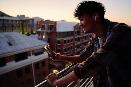 Foto de Vista del perfil del joven hombre multirracial en camisa sonriendo mientras usa el teléfono en el balcón del apartamento durante el atardecer - Imagen libre de derechos