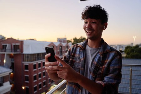 Foto de Joven hombre multirracial en camisa haciendo videollamada con teléfono móvil en el balcón del apartamento durante la puesta del sol - Imagen libre de derechos