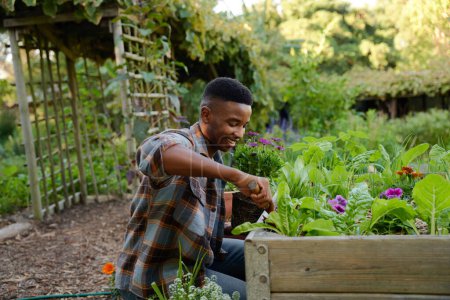 Foto de Happy young black man wearing checked shirt smiling while using tool in flowerbed in garden center - Imagen libre de derechos