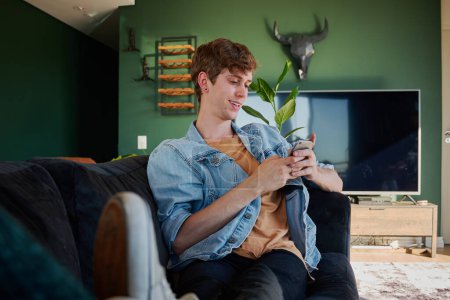 Foto de Happy young caucasian man in casual clothing sitting on sofa while using mobile phone in living room at home - Imagen libre de derechos