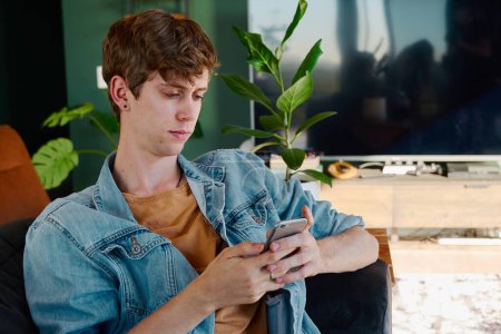 Téléchargez les photos : Young gay caucasian man in casual clothing using mobile phone on sofa in living room at home - en image libre de droit