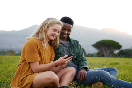 Foto de Young multiracial couple in casual clothing sitting and smiling while using mobile phone in natural parkland - Imagen libre de derechos