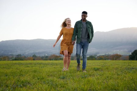 Foto de Young multiracial couple in casual clothing smiling and holding hands while walking in field - Imagen libre de derechos