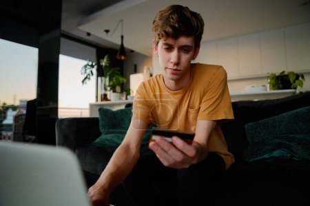 Téléchargez les photos : Young caucasian man in casual clothing holding credit card while using laptop in living room at home - en image libre de droit