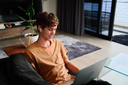 Téléchargez les photos : Young caucasian man in casual clothing working on laptop with wireless headphones on sofa at home - en image libre de droit