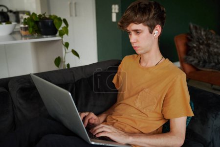 Photo for Young caucasian man wearing t-shirt working on laptop with wireless headphones on sofa at home - Royalty Free Image