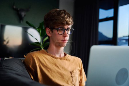 Foto de Young caucasian man wearing eyeglasses working on laptop in living room on sofa at home - Imagen libre de derechos