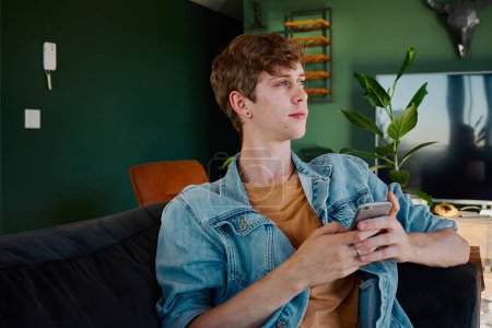 Téléchargez les photos : Young caucasian man in denim jacket looking away while using mobile phone in living room at home - en image libre de droit