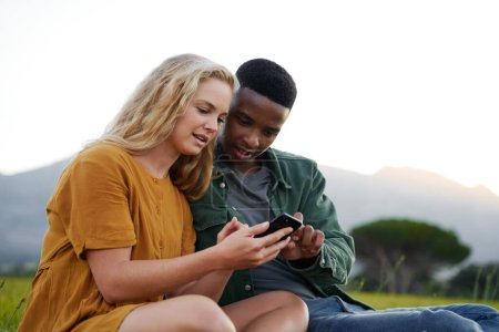 Téléchargez les photos : Young multiracial couple in casual clothing sitting and looking down while using mobile phone in field - en image libre de droit