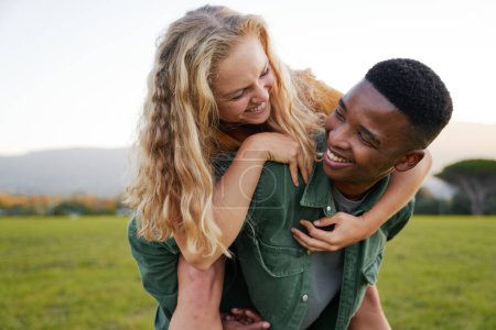 Foto de Happy young black man in shirt smiling while giving young caucasian woman piggyback in field - Imagen libre de derechos