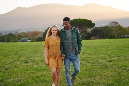 Photo for Happy multiracial young couple in casual clothing smiling and holding hands while walking in field - Royalty Free Image
