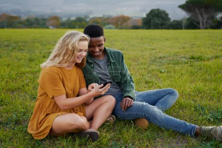 Téléchargez les photos : Happy multiracial young couple in casual clothing sitting and smiling while using mobile phone in field - en image libre de droit