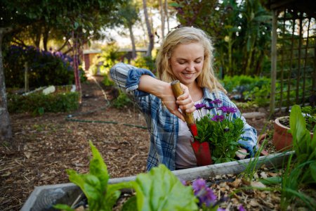 Foto de Happy young caucasian woman in checked shirt using spade while planting flowers in garden center - Imagen libre de derechos