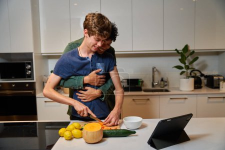 Foto de Young gay couple in casual clothing embracing while cutting vegetables by digital tablet in kitchen at home - Imagen libre de derechos