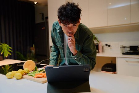 Photo for Young multiracial man in long sleeved shirt preparing dinner while checking digital tablet in kitchen at home - Royalty Free Image