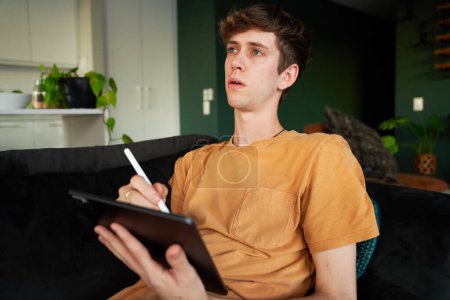 Téléchargez les photos : Young caucasian man in casual clothing using digitized pen with digital tablet in living room at home - en image libre de droit