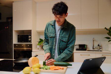 Foto de Young multiracial man in long sleeved shirt cutting vegetables next to digital tablet in kitchen at home - Imagen libre de derechos