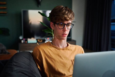 Téléchargez les photos : Young caucasian man in casual clothing and eyeglasses working on laptop in living room on sofa at home - en image libre de droit