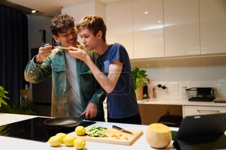 Foto de Young gay couple in casual clothing smiling while tasting dinner by digital tablet in kitchen at home - Imagen libre de derechos