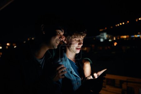 Foto de Happy young gay couple in casual clothing using mobile phone on balcony of apartment at night - Imagen libre de derechos