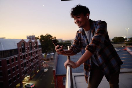 Photo for Young multiracial man in checkered shirt smiling while using phone on balcony of apartment during sunset - Royalty Free Image