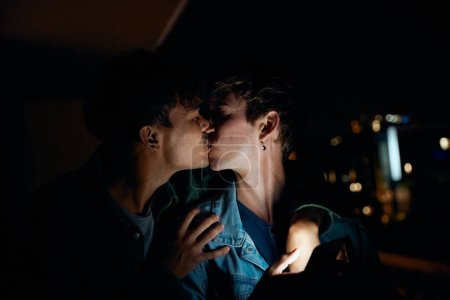 Photo for Young gay couple in casual clothing kissing while holding mobile phone on balcony of apartment at night - Royalty Free Image