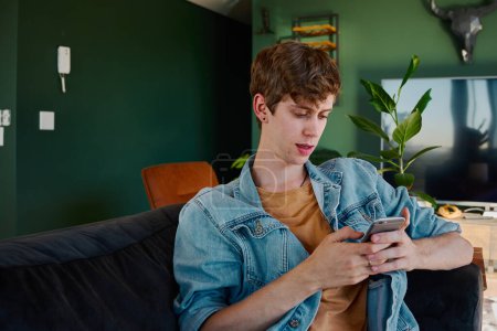 Téléchargez les photos : Young caucasian man in denim jacket looking down while using mobile phone in living room at home - en image libre de droit