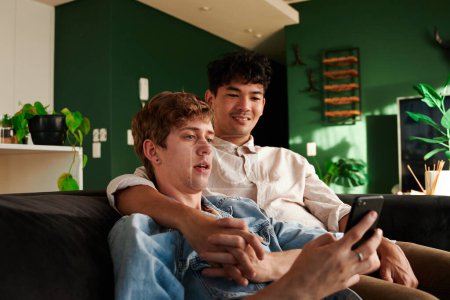 Foto de Happy young gay couple looking at mobile phone while holding hands on sofa in living room at home - Imagen libre de derechos